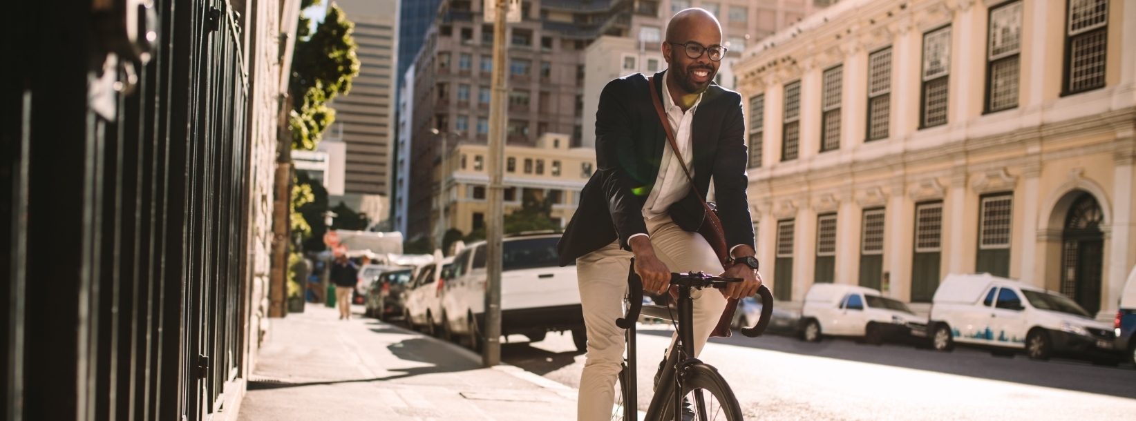 Banner image of A professionally dressed man rides a bicycle through a vibrant city street, smiling as he enjoys the morning sunlight. The urban setting, lined with historic buildings and bustling with activity, highlights a dynamic lifestyle that blends work and well-being, emphasizing mobility, sustainability, and the joy of commuting in an energetic metropolis.
