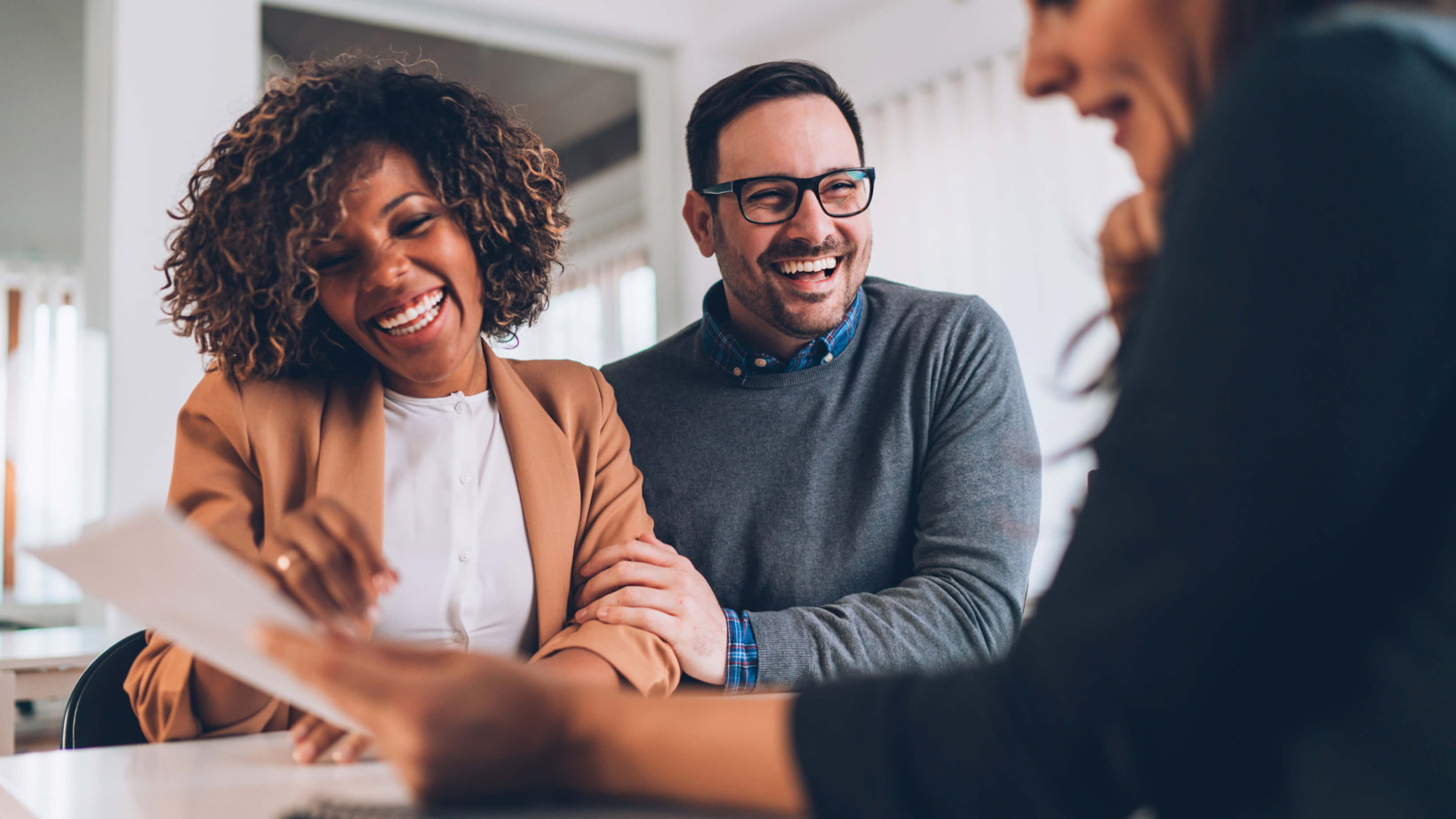 The banner image shows a happy couple sitting across from a professional, possibly a real estate agent or financial advisor, in a bright office setting. The couple is smiling and looking at a document that the professional is presenting to them. The woman is dressed in a stylish brown blazer, while the man wears glasses and a gray sweater. The professional, whose face is partially visible, is engaging with them in conversation. The image conveys a positive and professional atmosphere, possibly related to buying a home, signing a contract, or making an important financial decision.