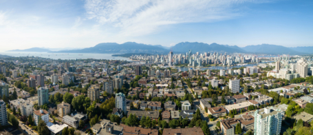Banner of panoramic view of Vancouver with towering buildings surrounded by lush greenery. Beyond the urban area lies a serene waterfront with calm waters. Majestic mountains rise in the background under a clear blue sky with wispy clouds, creating a breathtaking scene.