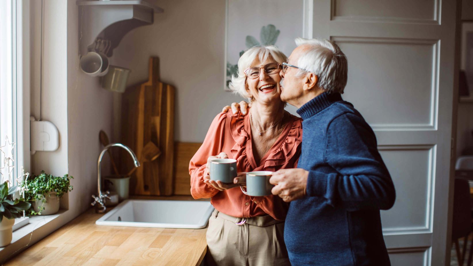 Picture of a happy elderly couple shares a warm moment in their cozy kitchen, holding coffee mugs while embracing. Their smiles reflect love, comfort, and the joy of a life well-lived together at home.