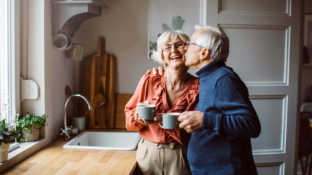 Picture of a happy elderly couple shares a warm moment in their cozy kitchen, holding coffee mugs while embracing. Their smiles reflect love, comfort, and the joy of a life well-lived together at home.