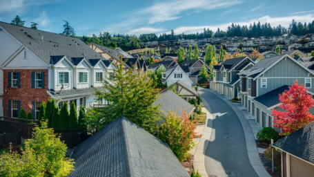 Banner shows a charming suburban neighborhood features neatly lined houses along a winding street. Lush greenery and vibrant autumn trees enhance the scene, with distant homes nestled against a backdrop of tall evergreens and a clear blue sky.