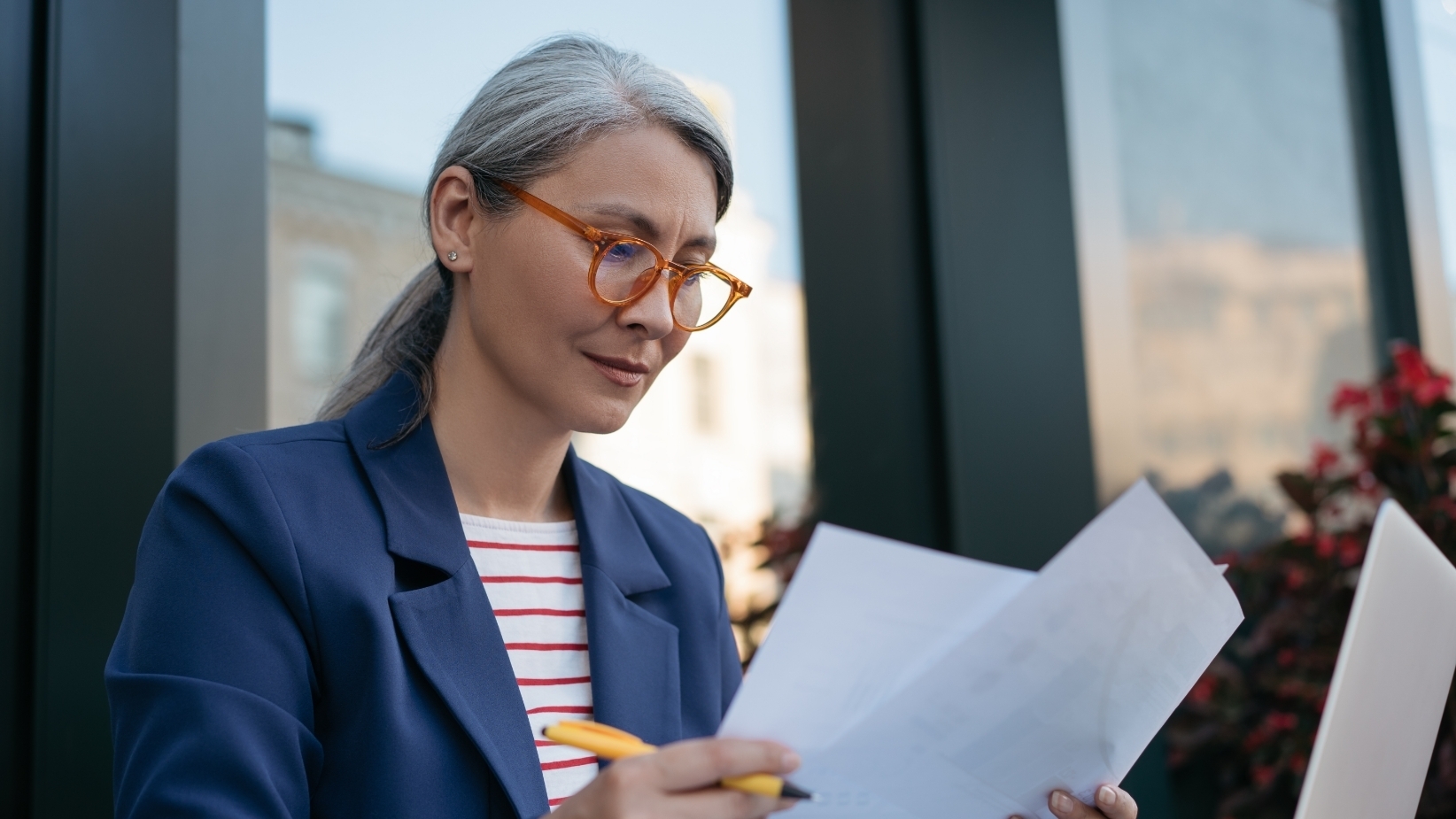 The banner image shows a professional woman with gray hair wearing orange-framed glasses and a blue blazer over a striped shirt. She is seated outdoors, holding papers and a pen, appearing focused. The background includes a modern glass building and some plants.