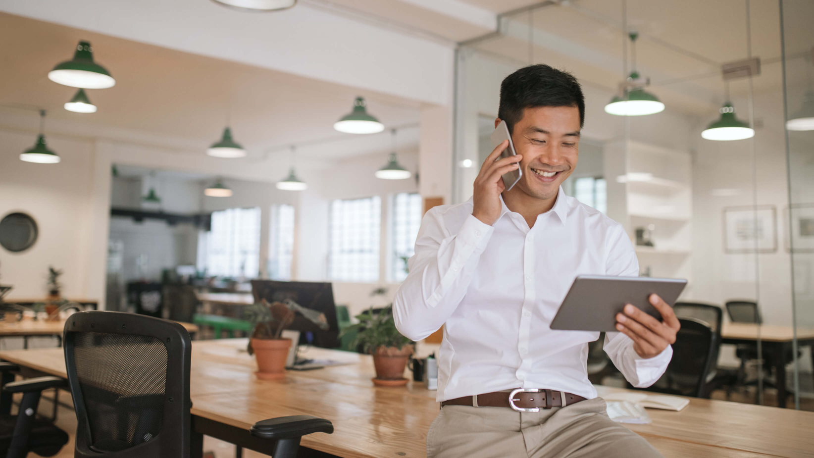 The banner shows a professional man in a modern office space smiles while talking on his phone and reviewing a tablet. The bright, open workspace with glass partitions and stylish lighting reflects a dynamic, collaborative environment, ideal for productivity and innovation.