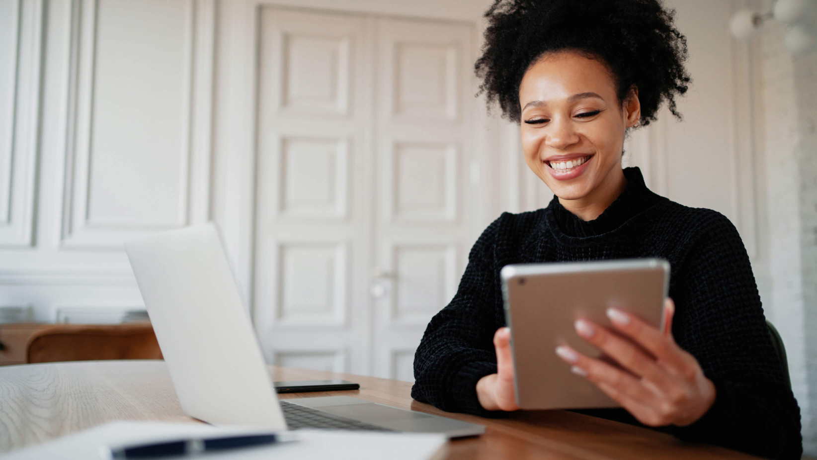 Banner showing a smiling woman sitting at a desk, holding a tablet while working. A laptop, notebook, and pen are on the table. She appears engaged and happy in a bright, modern room with white paneled doors.