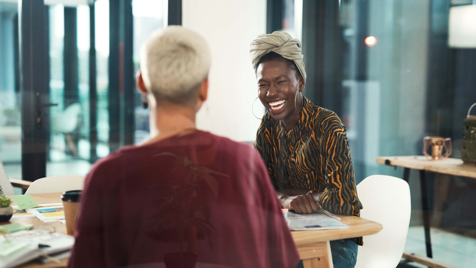 The banner image captures two women engaged in a lively conversation at a modern, well-lit workspace. One woman, wearing a headwrap and patterned blouse, is laughing joyfully, while the other, with short platinum hair, listens attentively. The setting features warm lighting, coffee cups, and paperwork, creating a welcoming and collaborative atmosphere.