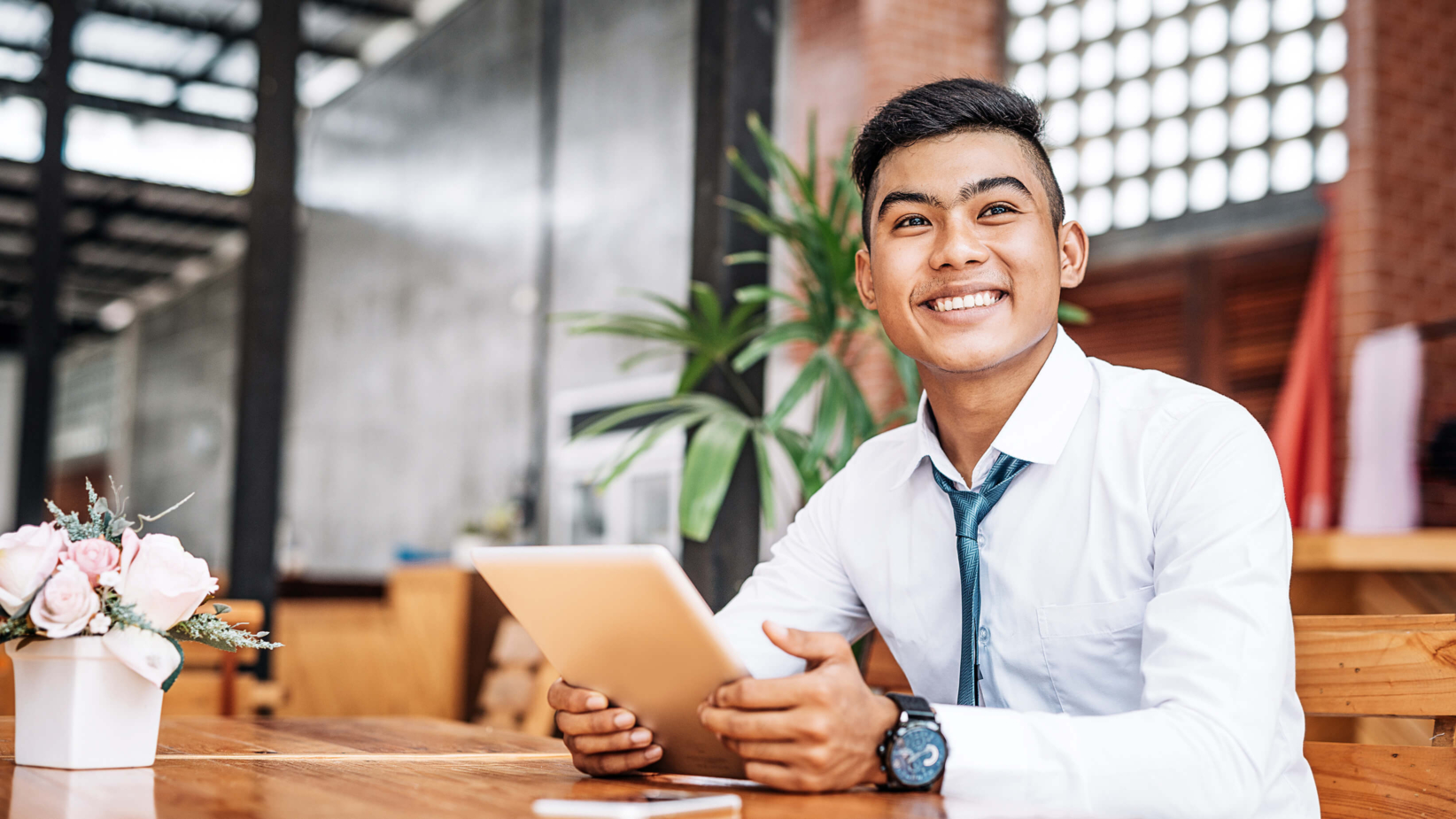 The image features a young man in a white shirt sitting in a modern workspace. He is smiling confidently, with a relaxed posture. The background includes bookshelves, greenery, and a casual office setting, creating an inviting and productive atmosphere.