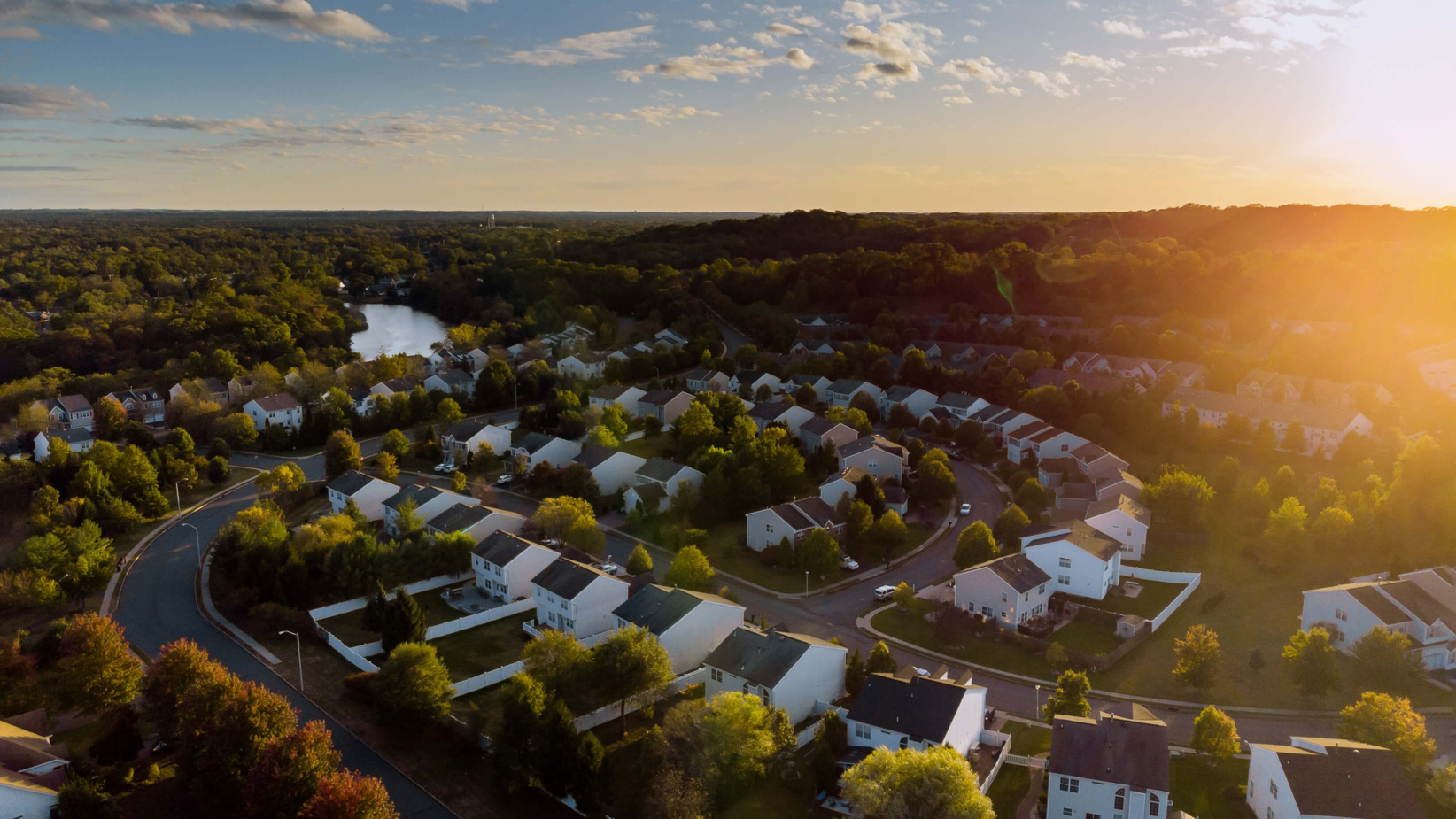 Banner shows an aerial view of a peaceful suburban neighborhood at sunset shows neatly arranged houses with tree-lined streets. The golden sunlight bathes the rooftops and surrounding greenery, creating a warm and tranquil atmosphere. A serene lake and dense forest stretch into the distance, enhancing the scenic beauty.