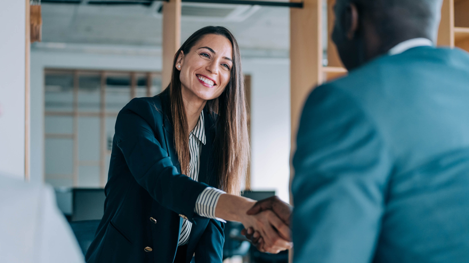 banner image of A professional woman in a business suit warmly shakes hands with a colleague in a modern office setting. Her confident smile and engaging demeanor reflect a positive and welcoming atmosphere, emphasizing professionalism, partnership, and successful business relationships.