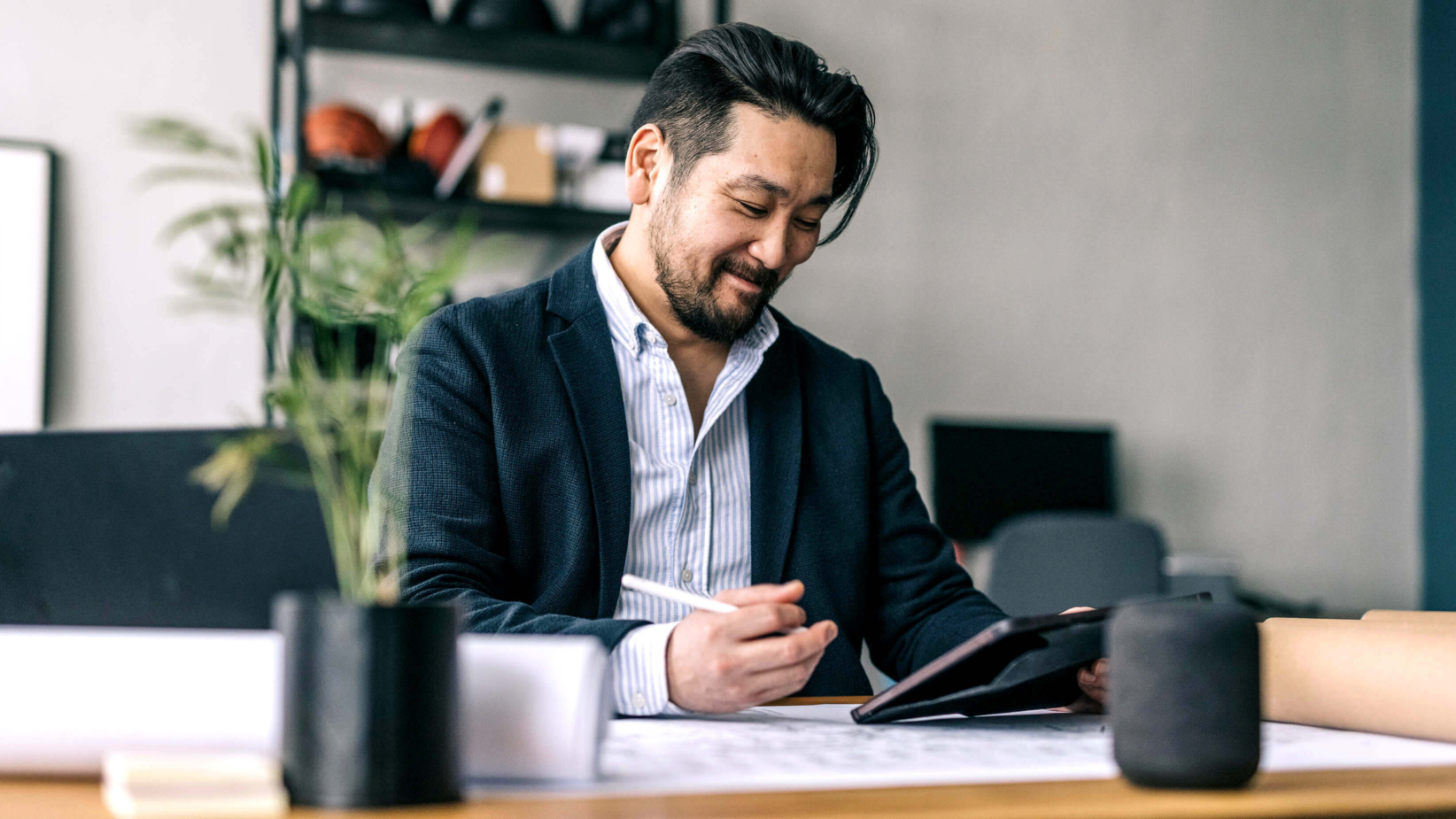 Banner shows a man in a business suit smiles while working at his desk, holding a stylus and tablet. Surrounded by office supplies, a potted plant, and rolled papers, he appears focused and content in a modern workspace.