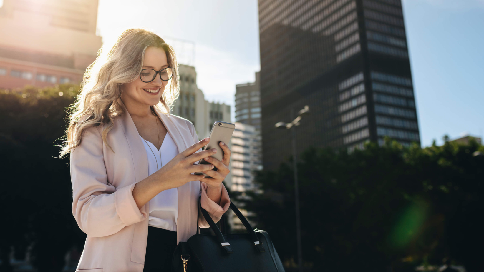 The banner image shows a professional woman standing outdoors in a cityscape, holding her phone and smiling. She is wearing a light pink blazer, glasses, and carries a black handbag. The background features tall office buildings, greenery, and sunlight creating a warm and vibrant atmosphere.