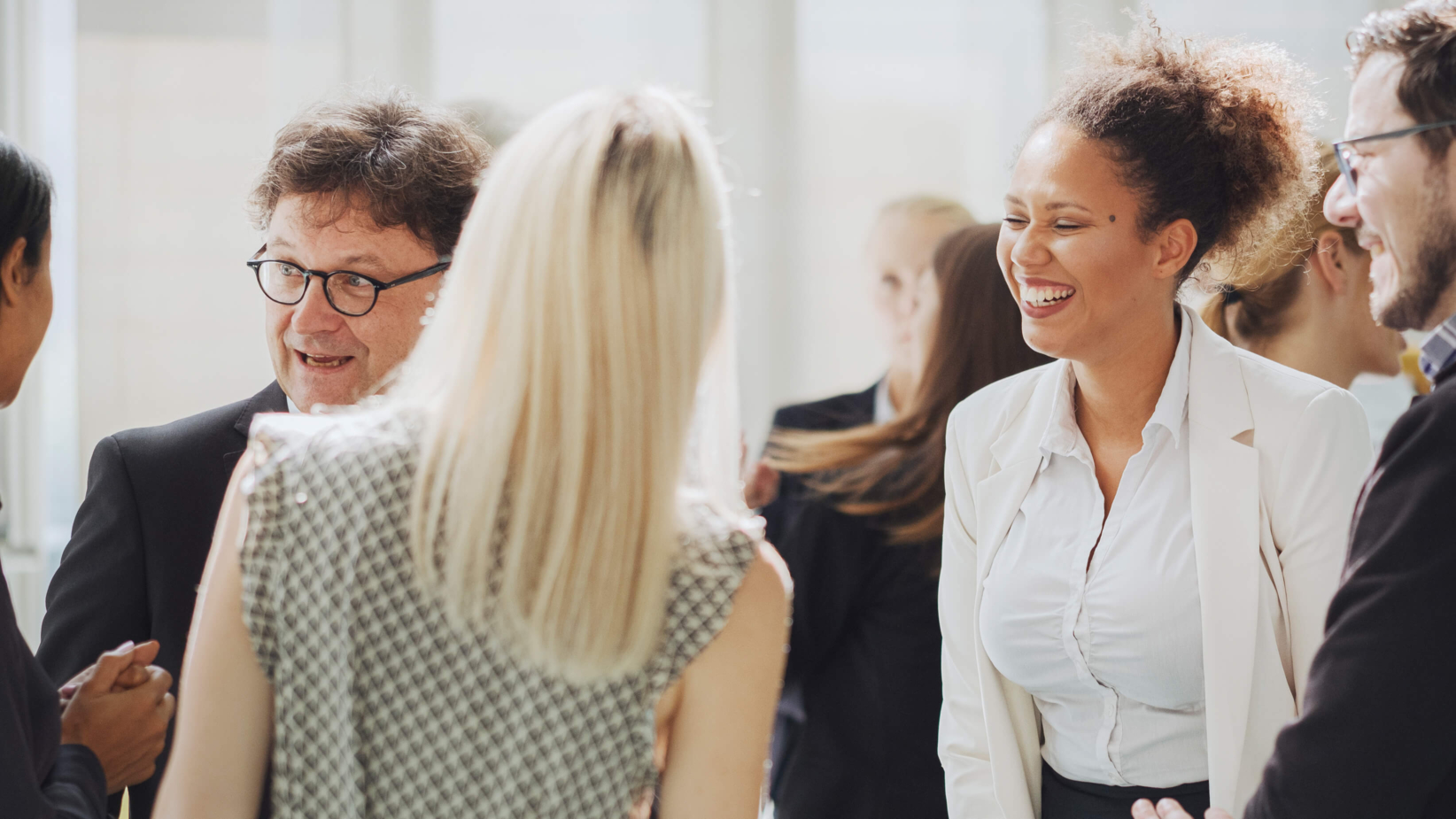 The banner image showcases a group of professionals engaged in lively conversation at a networking event. A woman in a white blazer is laughing, while others around her exchange smiles and handshakes. The bright, modern setting and the participants' enthusiastic expressions convey a sense of collaboration, connection, and community in a professional environment.