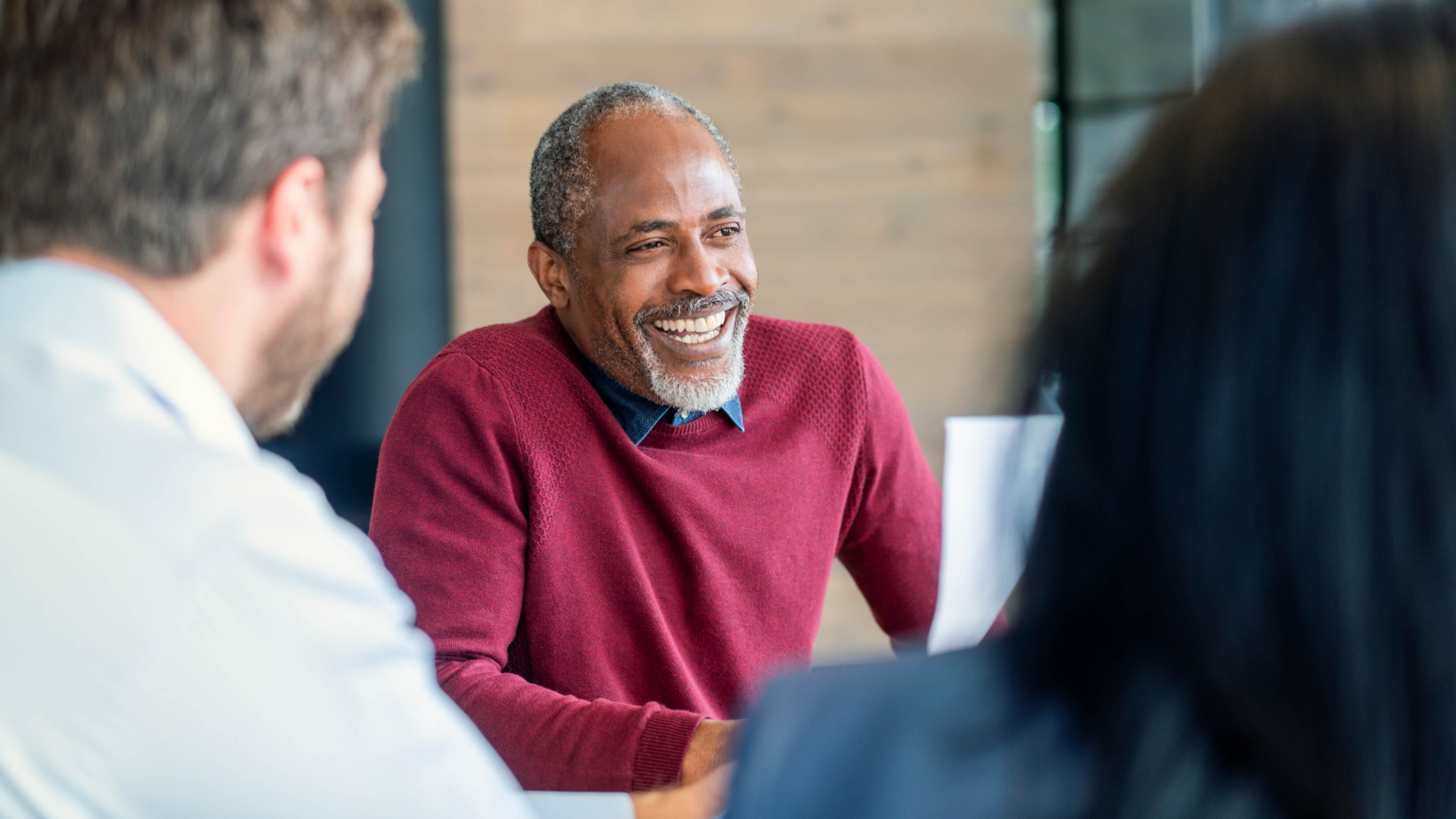 Images shows a confident and approachable leader engages in a discussion, exuding warmth and expertise. His genuine smile fosters a collaborative atmosphere, encouraging open communication. The professional setting, with colleagues attentively listening, reflects teamwork, leadership, and a commitment to shared success. This moment captures the essence of effective decision-making and mentorship.