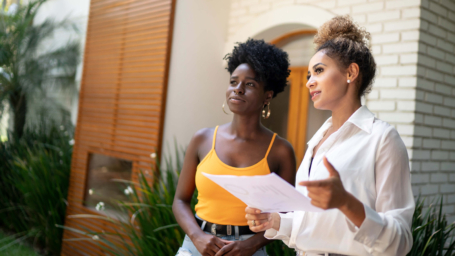 The banner shows a professional real estate agent, dressed in a white blouse, discusses property details with a potential homebuyer in a stylish outdoor setting. Holding documents, she gestures confidently while the client listens attentively, reflecting engagement in the home-buying process.