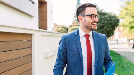 The banner image shows a professional man in a blue suit, red tie, and glasses walking outdoors with a smile on his face. He is holding a blue folder or document in his hand, suggesting he might be a real estate agent, business professional, or consultant. The background features a modern residential or commercial area with a white fence, wooden accents, greenery, and a sidewalk. The setting is bright and inviting, with natural sunlight adding a warm and positive atmosphere. The image conveys confidence, professionalism, and success.